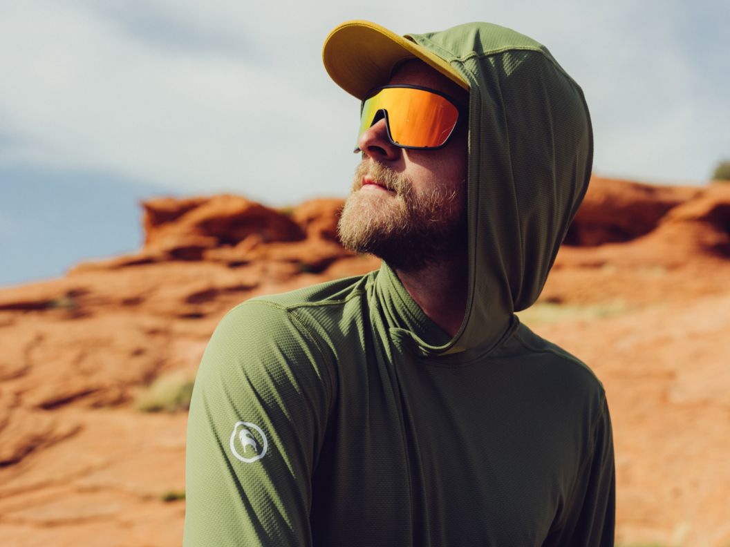 A bearded man wearing cool-guy sunglasses and a green sun hoodie in front of a red rock landscape.  