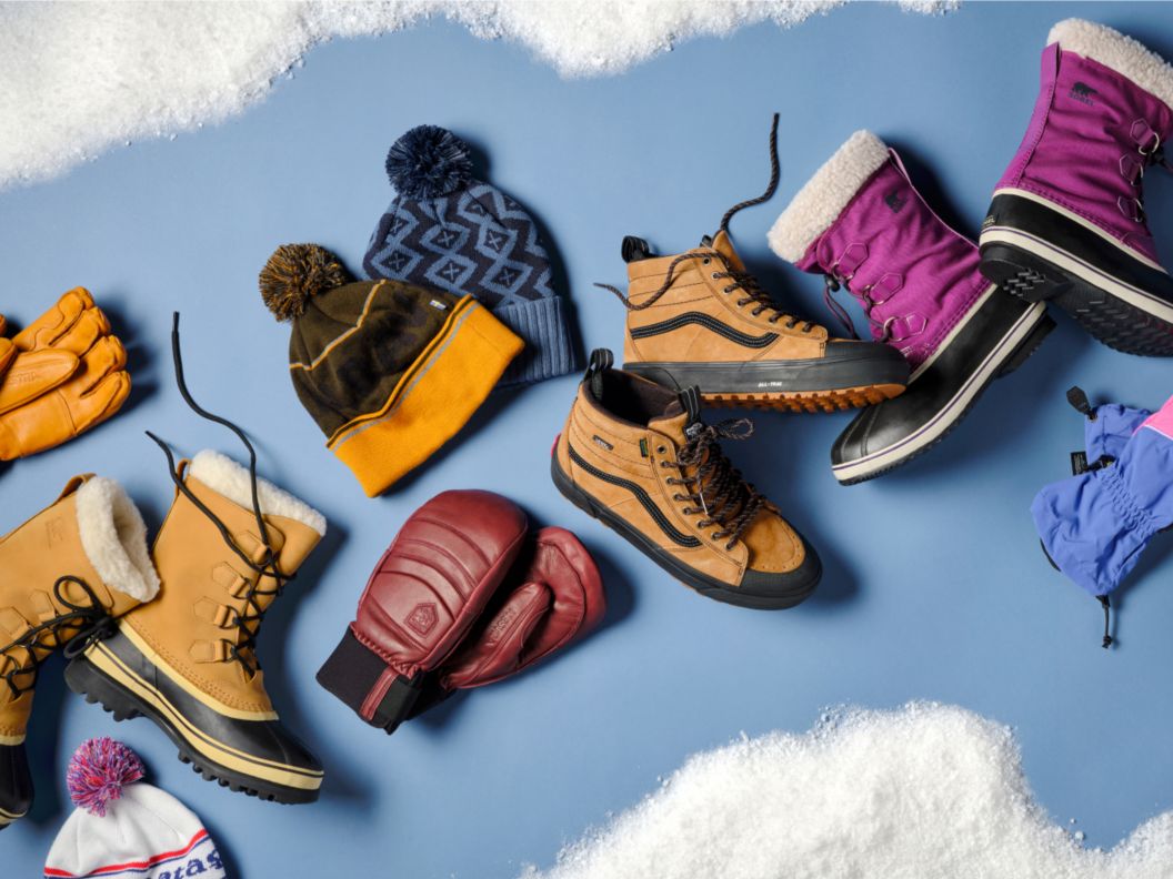 Snow boots, beanies, and mittens arranged on a blue backdrop, surrounded by faux snow.