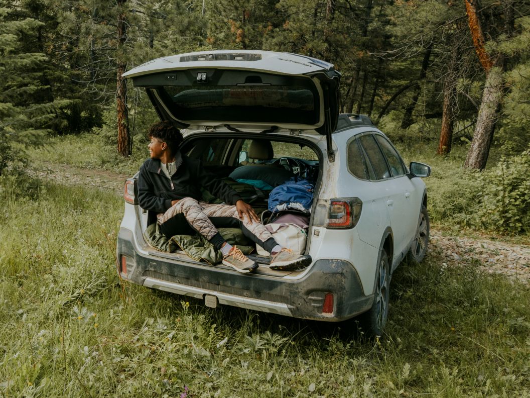 A smiling woman lays in the back of a packed car. The car is in a green field with trees around. She looks happy to be car camping.