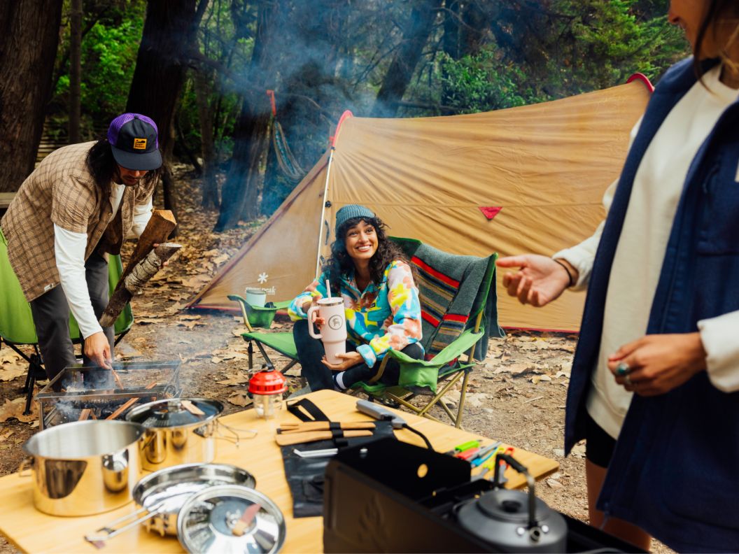 Someone prepares a camping meal as two people in camp chairs tend a fire in front of a large tent.