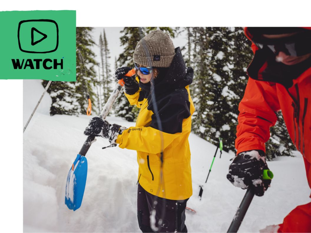 Two people dig a snow pit with backcountry shovels. A play button icon and the word “watch.” 