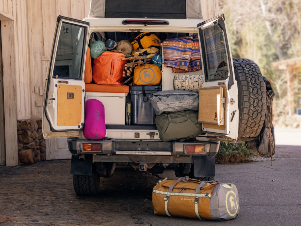 A gear-packed van has its back door open showing a cooler, blankets, and brightly colored bags.