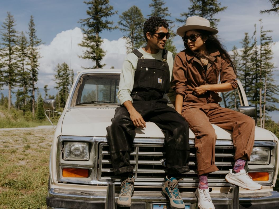 A man wearing black overalls and a woman in a brown jumpsuit sit on the hood of a vintage truck. 