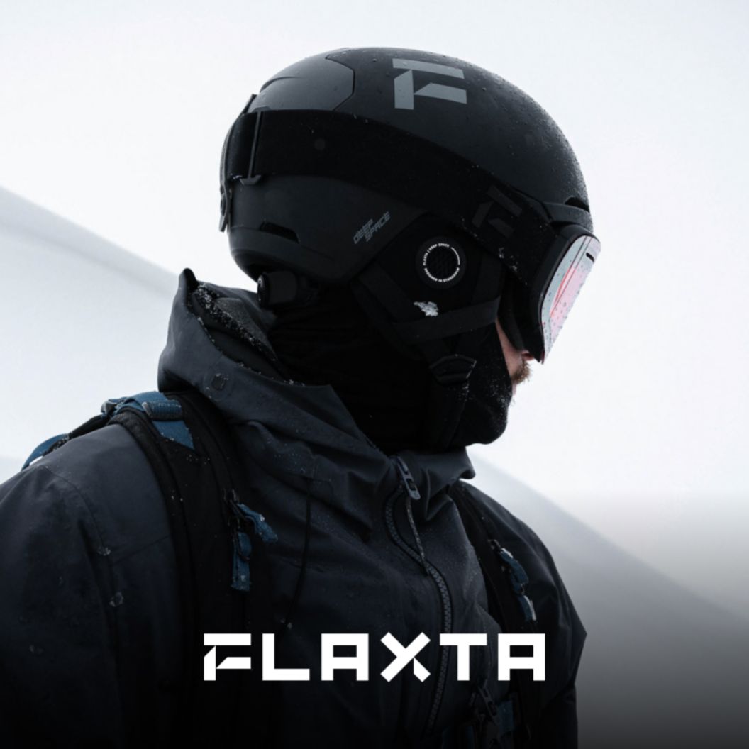 A person in a black snow kit, goggles, and helmet looks away from the camera. The Flaxta logo appears at the bottom of the image. 