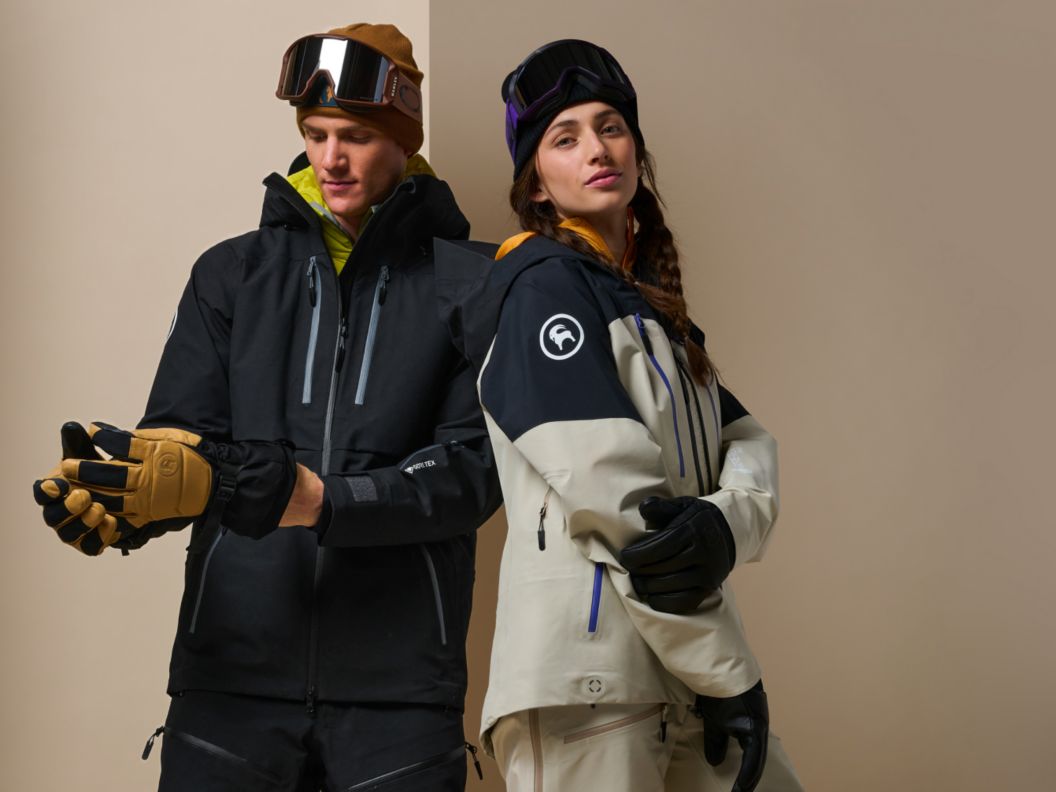Two models wearing an all-black outerwear kit and a cream and black colored outerwear kit against a tan background.