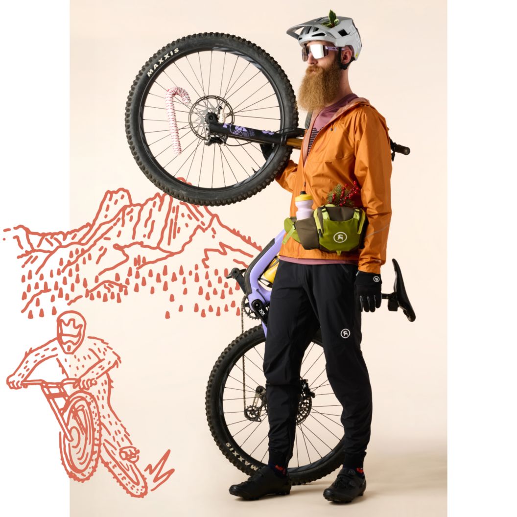 A mountain biker wearing a wind layer, riding pants, and a hip pack next to a mountain bike