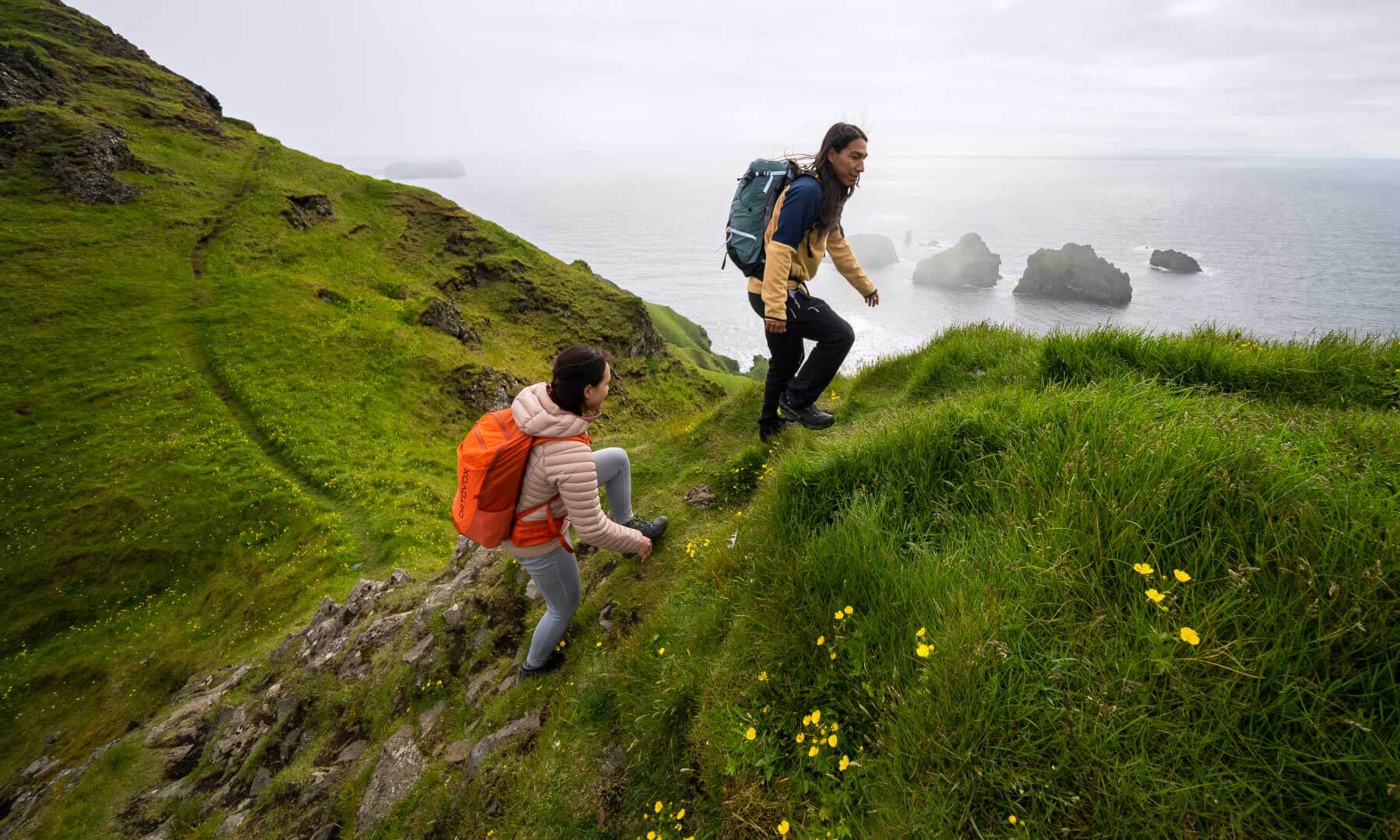 A man and woman hike on a narrow, grassy ridge overlooking an ocean with scattered islands.