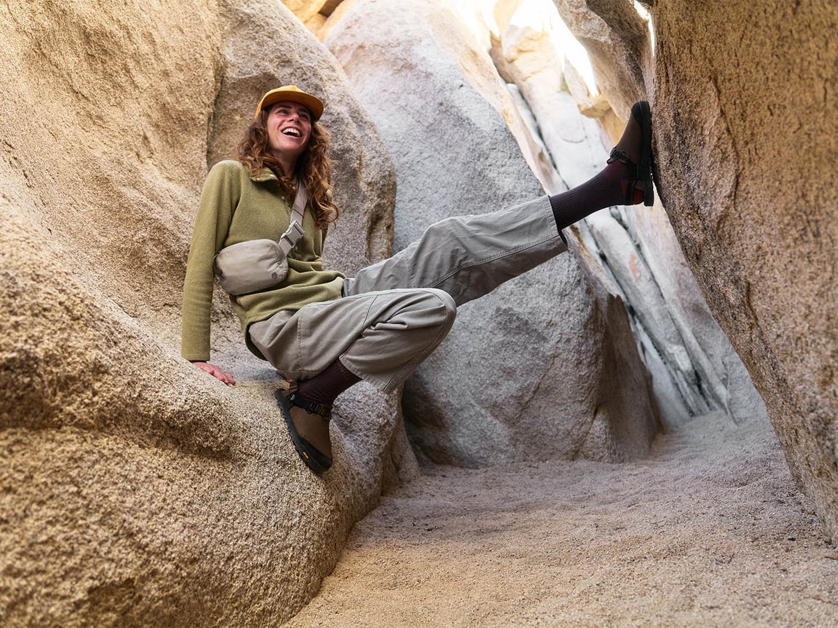 Smiling person in a narrow granite slot canyon with one leg braced against the rock wall, wearing dark Bedrock Mountain Clogs