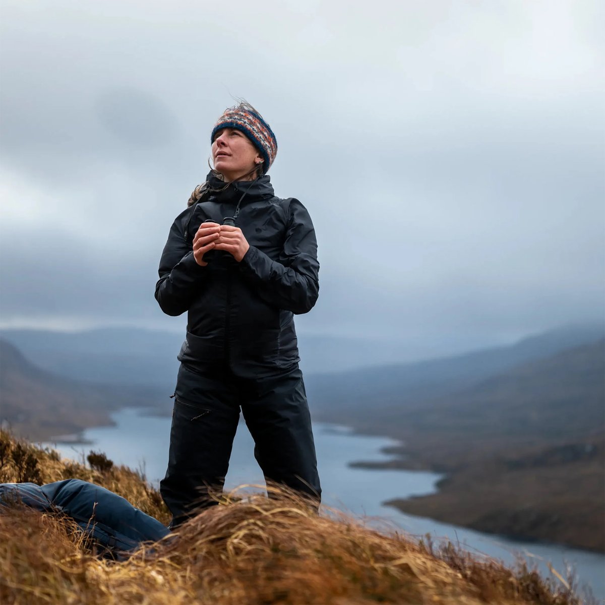 A woman stands on a windswept hill, wearing Fjallraven Keb trekking gear.