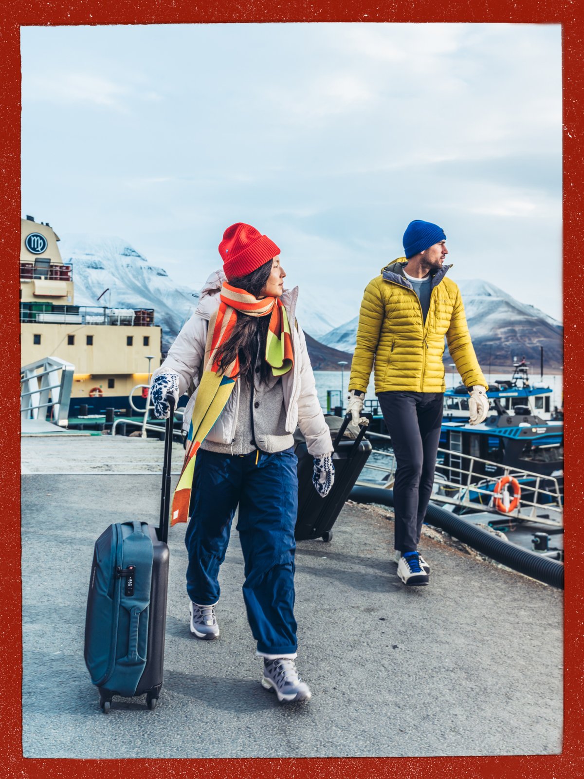 Two travelers pulling wheeled carry-on suitcases along a harbor dock with snowy mountains behind them.