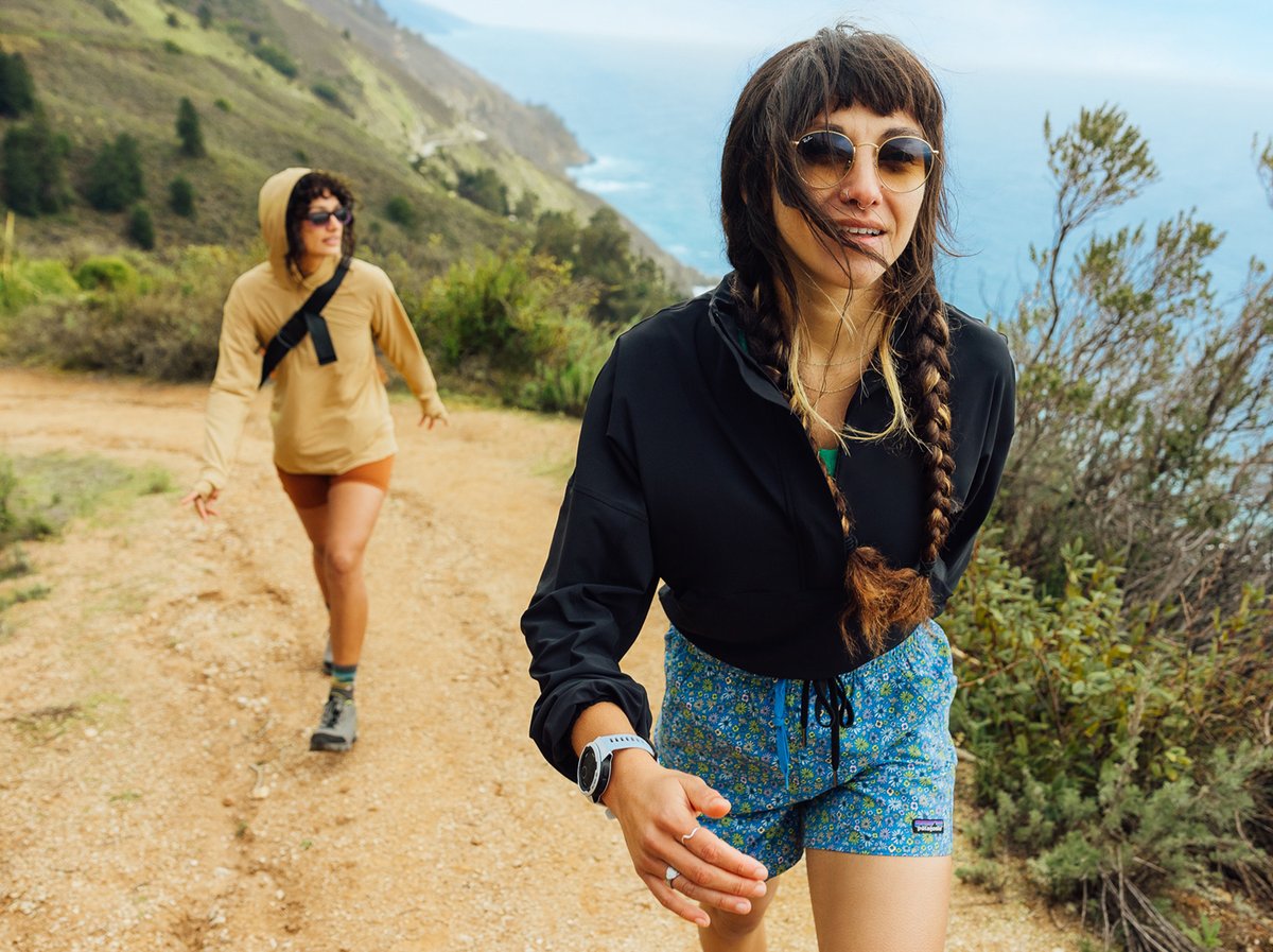 Two people hike on a dirt trail along a coastal hillside, with greenery, trees, and the ocean visible in the background. The person in the foreground wears a black hoodie, blue patterned Patagonia Baggie shorts, and a watch, while the person behind wears a  tan Tahoe sun hoodie, shorts, and hiking boots.