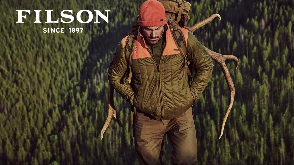 A man wearing an orange-paneled jacket and an orange beanie carries large elk antlers on his back through a green forest.