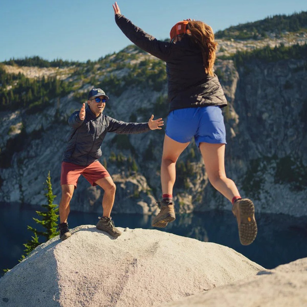 A man stands on a boulder, arms extended to catch a woman who is jumping across a gap in the boulder.