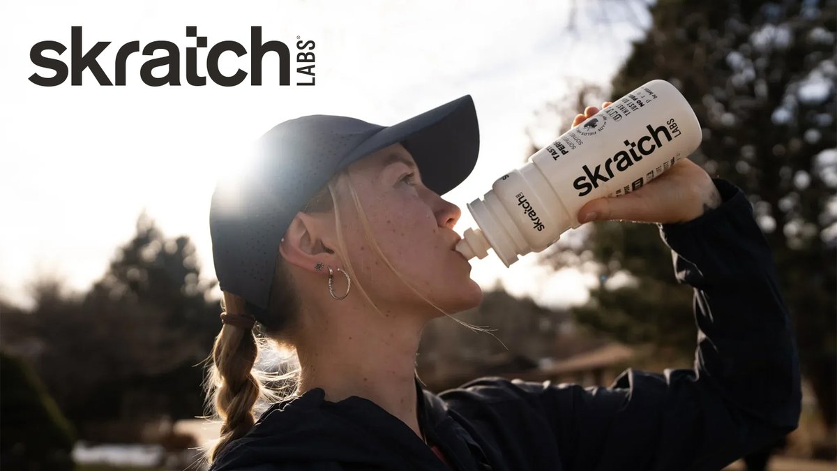 Woman drinking from a Skratch Labs hydration bottle outdoors while wearing a black cap on a sunny day with trees in the background.