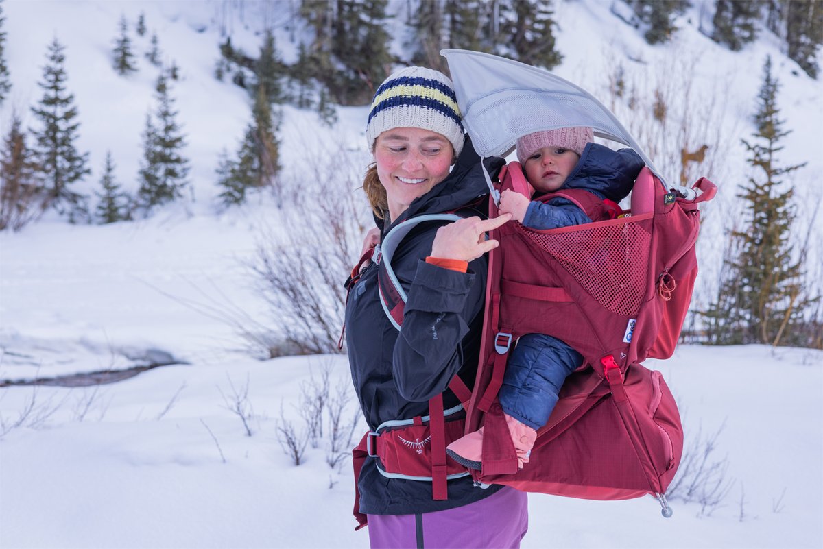 A smiling woman carries a child in a red Osprey carrier in the snow.