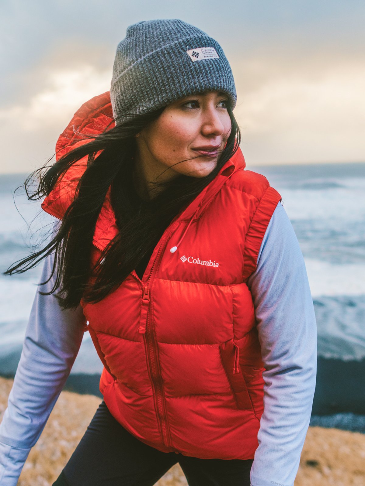 A woman wearing a bright red Columbia puffer vest over a long-sleeve shirt gazes at the waves on a cold day.