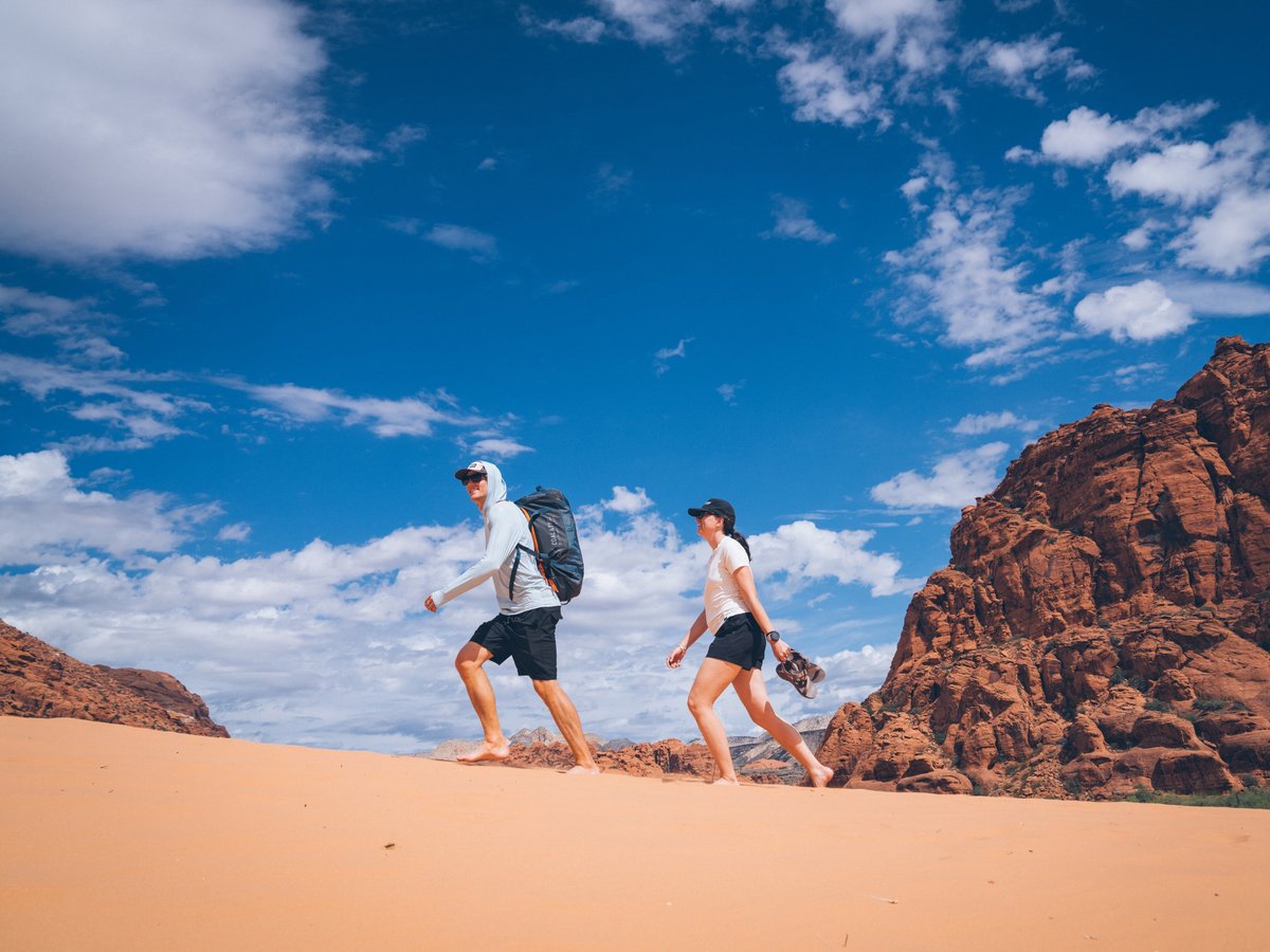 Two hikers walking barefoot across a red sand dune with sandstone buttes and a blue sky behind them.