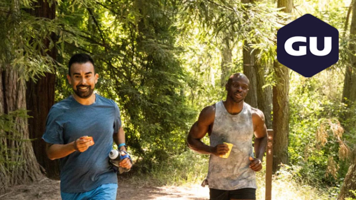 Two male runners jogging together on a tree-lined forest trail holding GU energy products and wearing athletic apparel during outdoor running exercise.