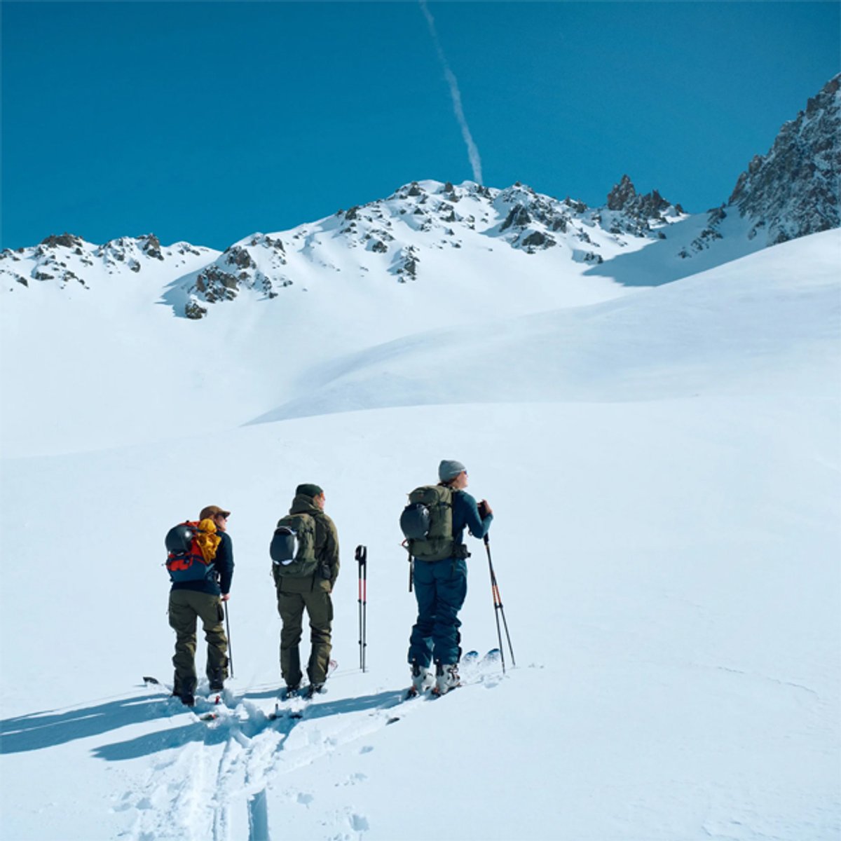 Three skiers in winter gear stand on a snowy mountain slope with clear blue skies and rocky peaks in the background during a backcountry skiing adventure.