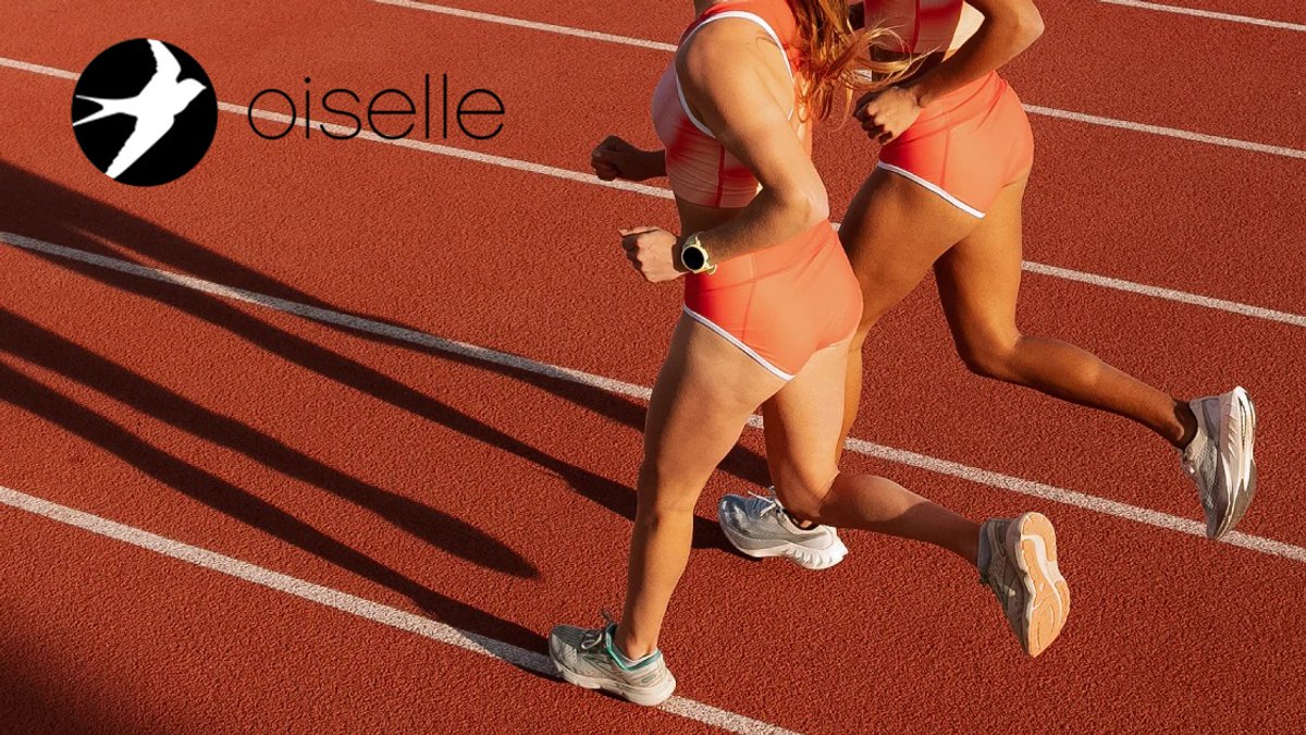 Two female runners in coral athletic wear jogging on a red running track with lane markings visible.