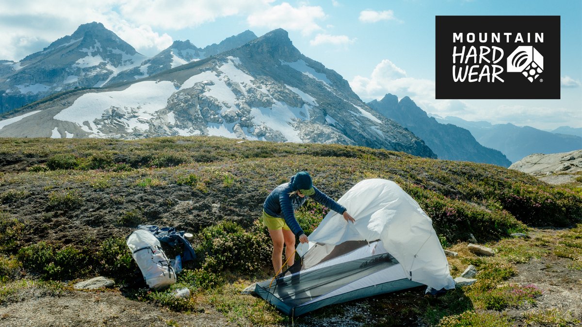 A camper sets up a white Mountain Hardwear tent in a high mountain meadow with snow-capped peaks in the distance.