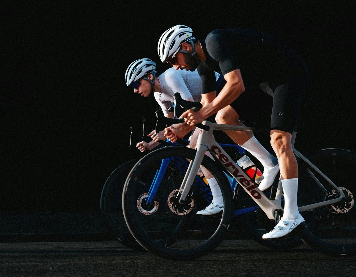 Two road bike riders in aggressive aero position drafting on Cervélo carbon road bikes against dark dramatic background
