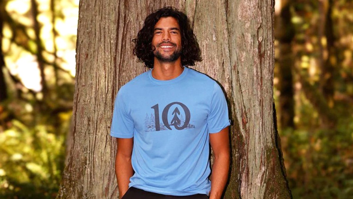 Man wearing light blue t-shirt with Tentree logo standing against large tree trunk in forest setting.