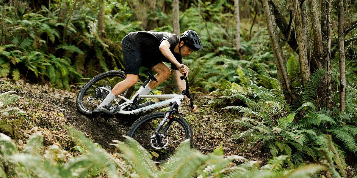 Rider in a black kit and helmet cornering a white full-suspension mountain bike through a fern-lined muddy forest trail.