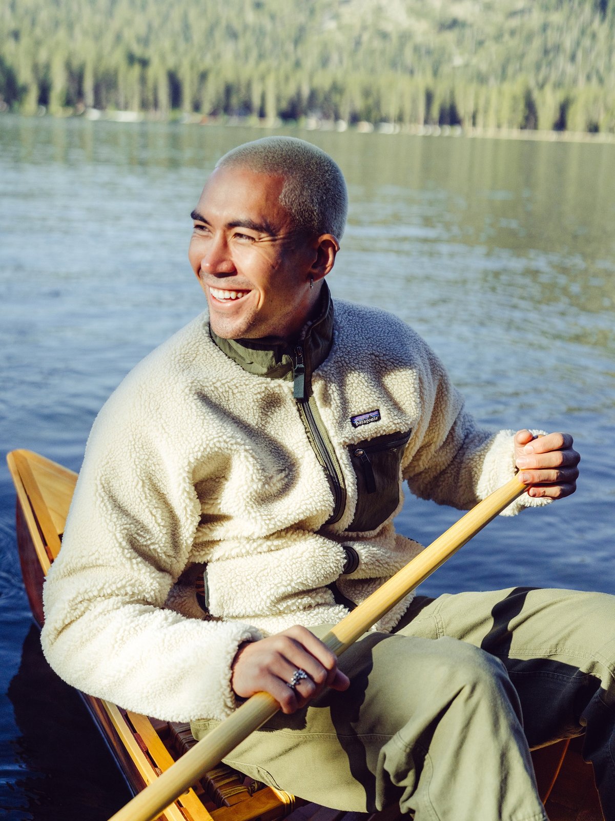 A person wearing a light-colored fleece jacket and olive green pants paddles a canoe on a calm lake.