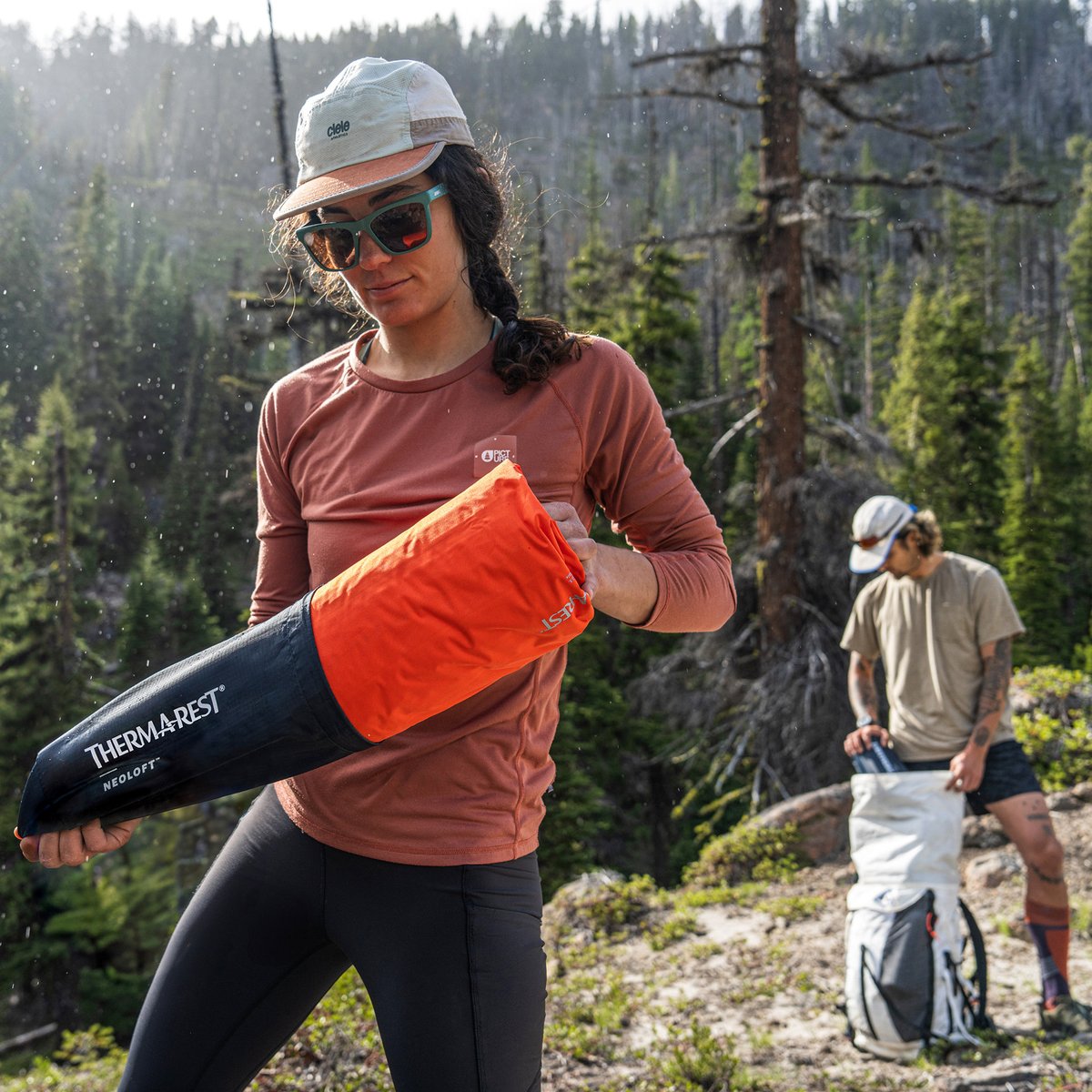 A woman in a remote forested locale removes her NeoLoft pad from its packsack. Behind her, a man removes gear from an ultralight backpacking pack.