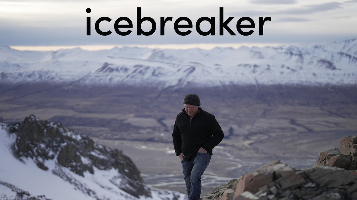 A man in a black quarter-zip sweater and beanie walks along a rocky mountain ridge with snow-covered peaks in the distance.