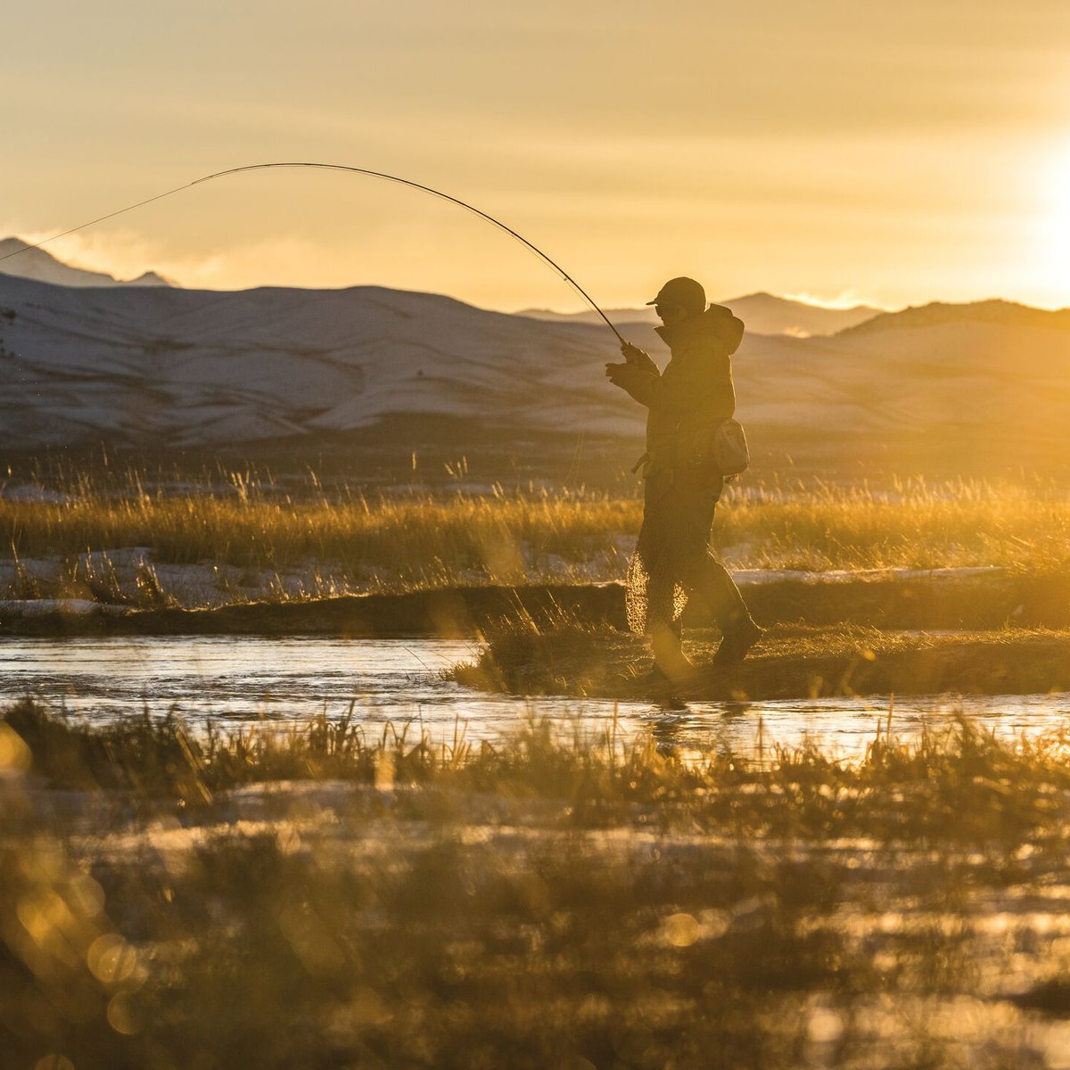 Fly fisherman casting a line in a shallow mountain stream at golden hour sunrise with scenic peaks and grassland in the background.