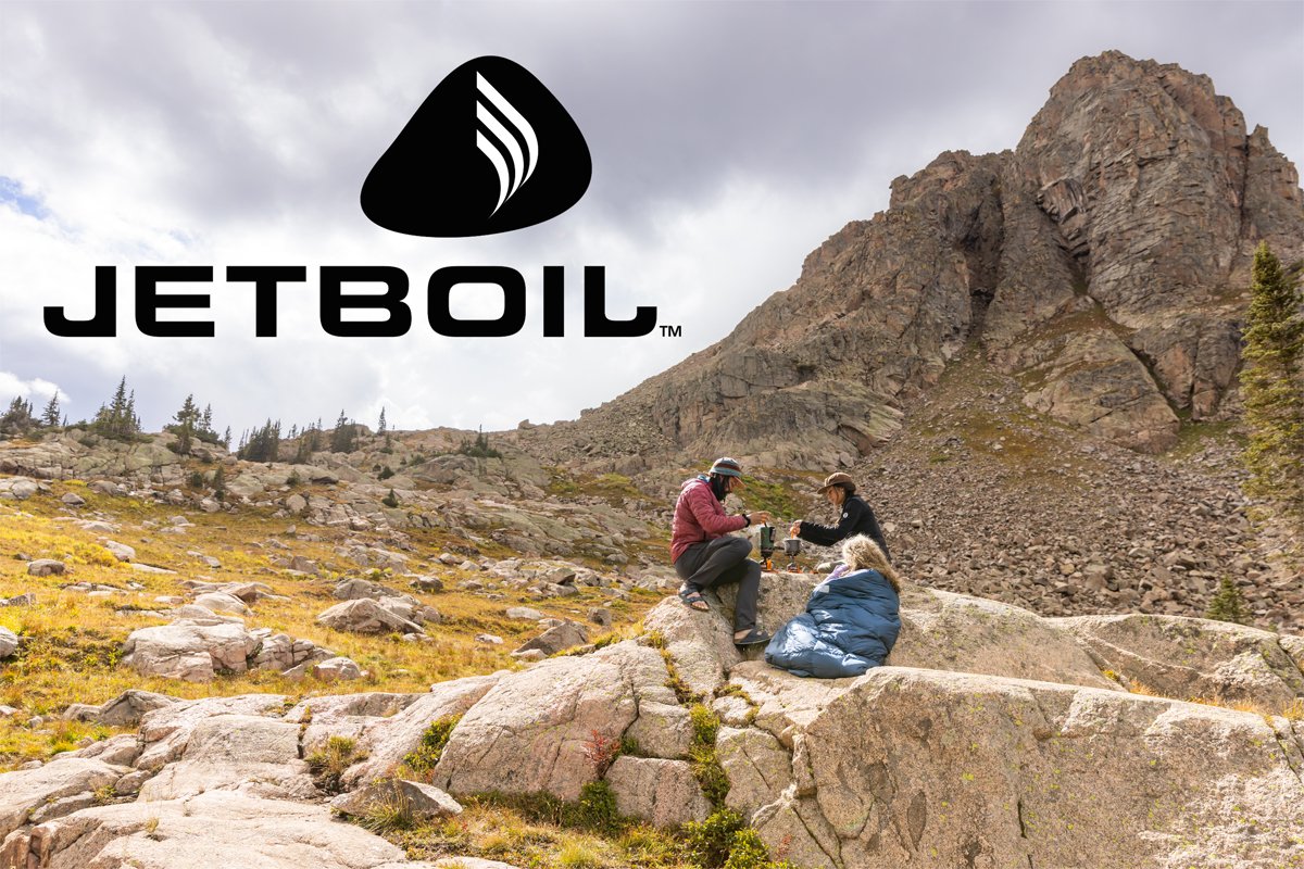 Three people sit on a boulder while cooking on a JetBoil stove.