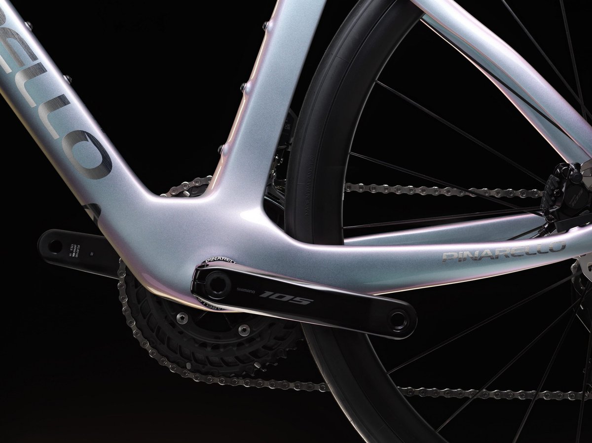 Pinarello road bike carbon bottom bracket and Shimano 105 crankset detail in iridescent silver-pink finish against dark studio background