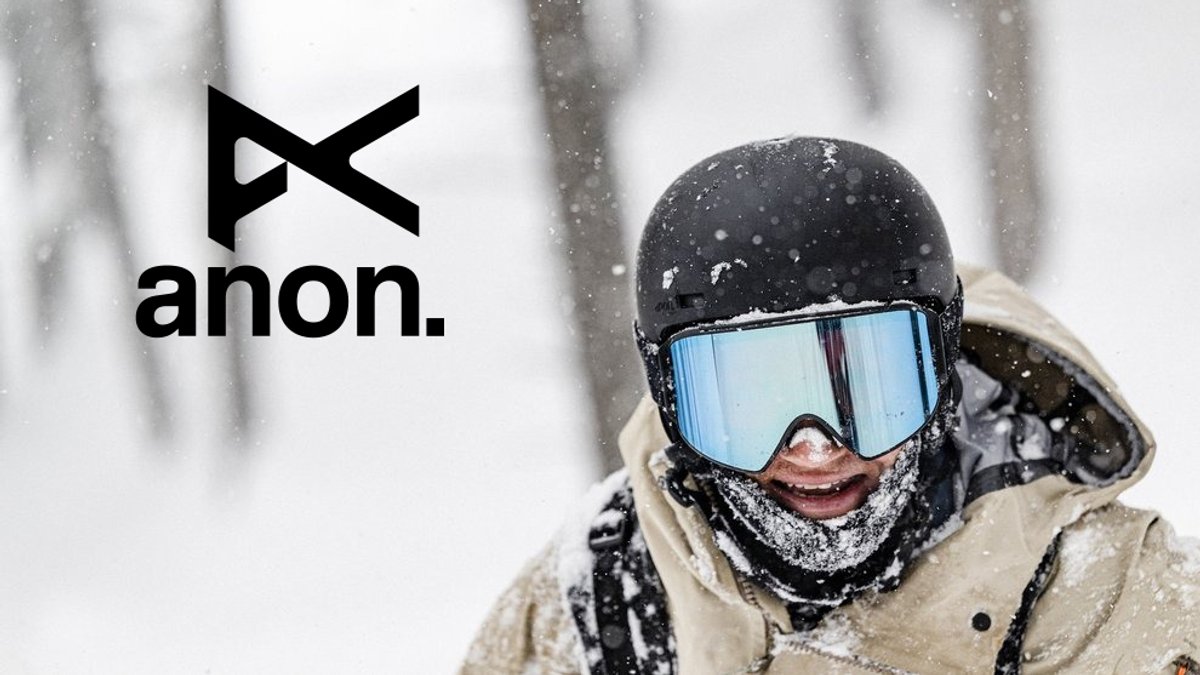A snowboarder wearing a tan jacket and blue-mirrored goggles smiles while snow falls, next to the Anon brand logo.