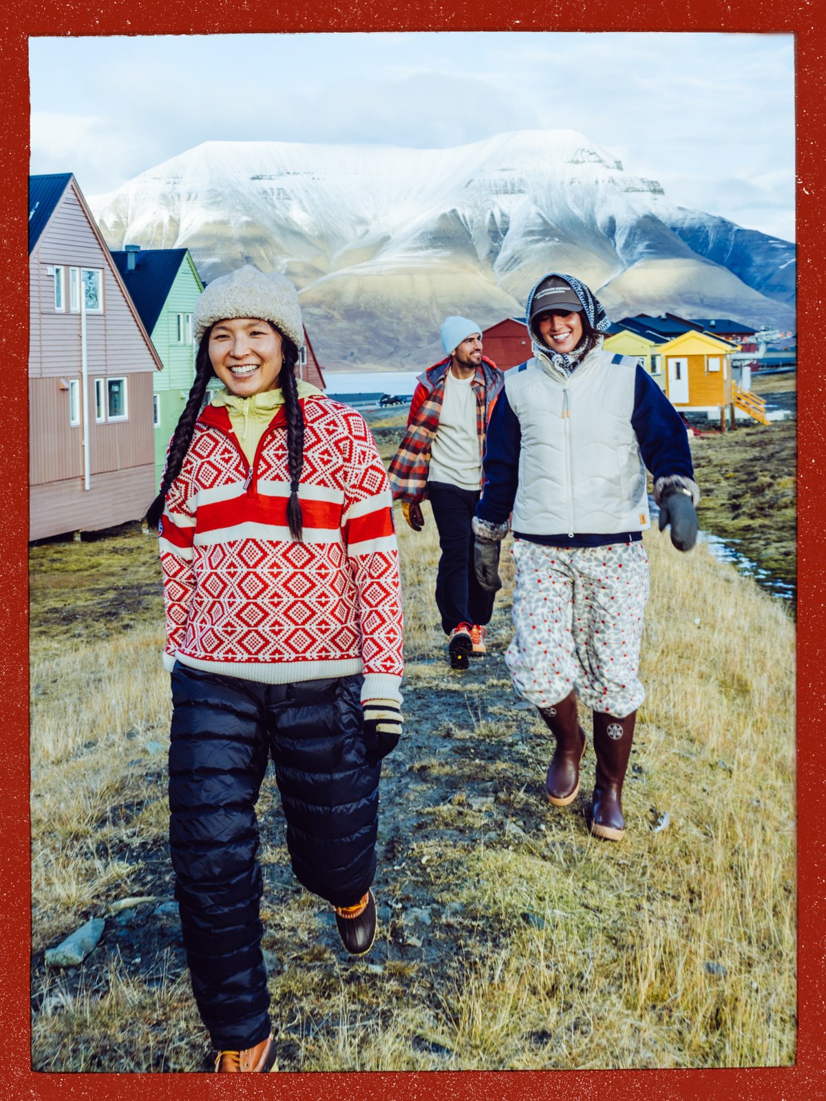 Three friends walking a grassy path past colorful wooden houses, with a snow-dusted mountain behind them.
