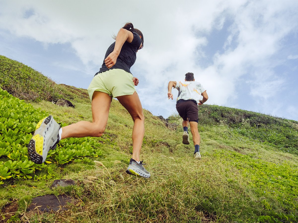 Two people running up a steep hillside trail with blue sky above and green hilly land below.