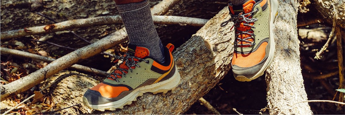 A person wearing Merrell Speed Solo hiking shoes with orange and olive-green accents stands on fallen tree branches in a wooded area.