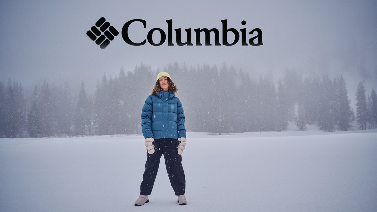 A woman in a blue Columbia puffer jacket and yellow beanie stands in a snowy field with a pine forest in the background.