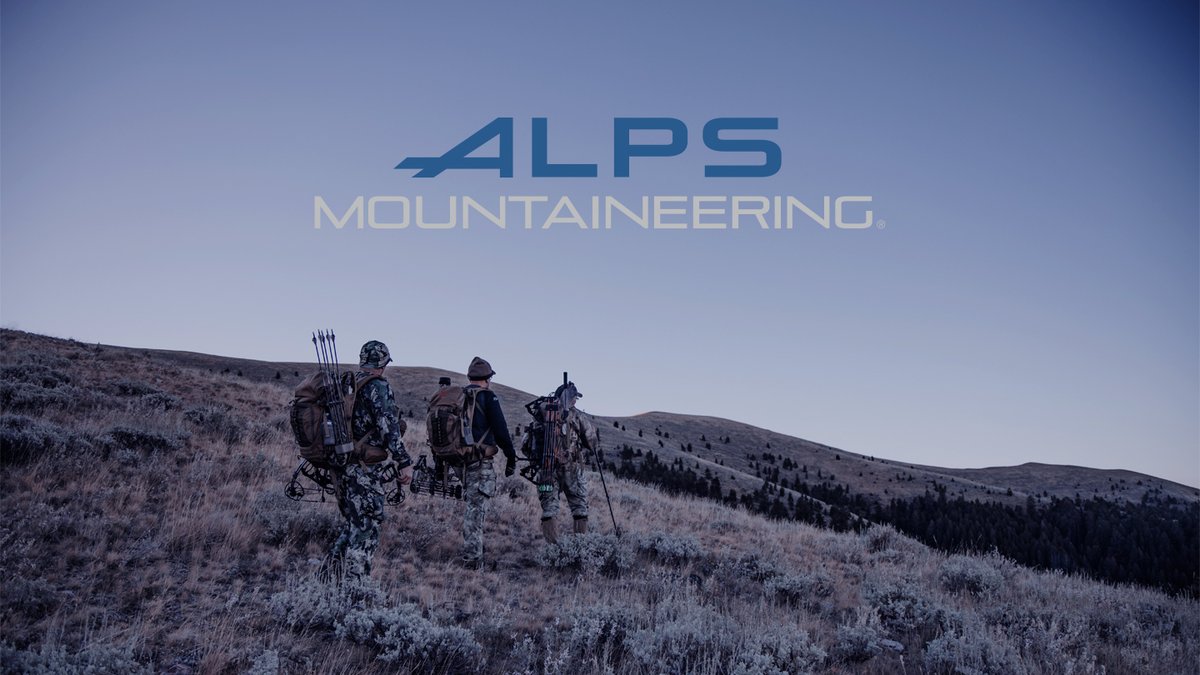 Four hunters in camouflage gear carrying bows and backpacks trek across sagebrush-covered hills under clear skies with ALPS Mountaineering branding.