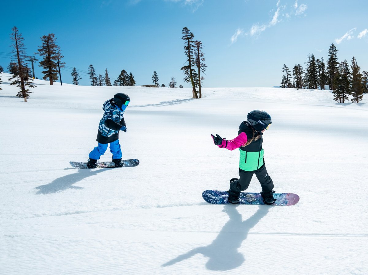 Two young kids snowboarding down a sunny groomed run surrounded by pine trees.