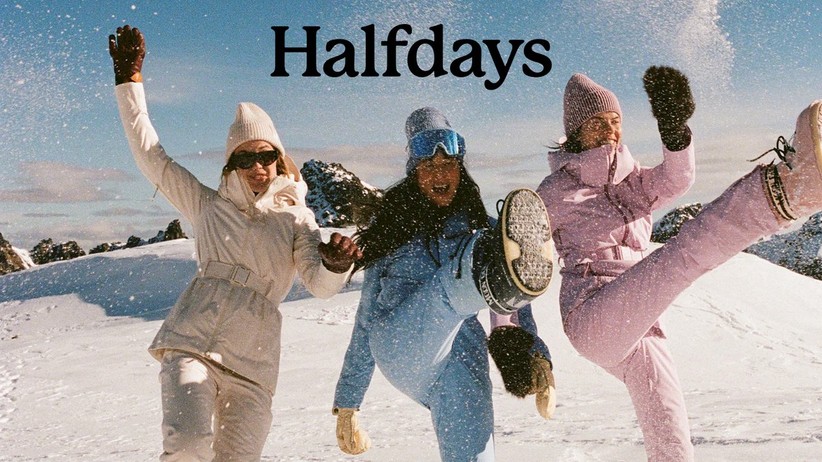 Four people in winter clothing jump joyfully in the snow with their arms raised, celebrating halfdays in a snowy mountain landscape.