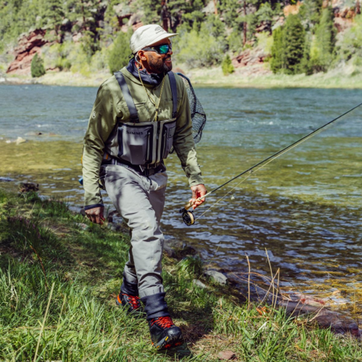 A fly fisherman wearing waders walks along a river bank.