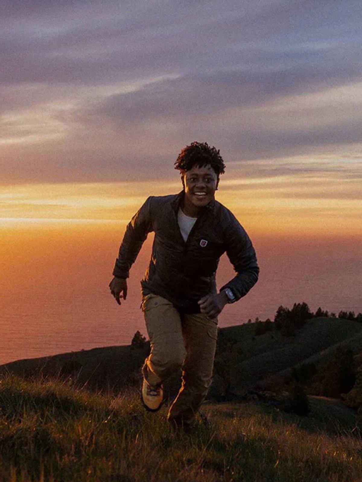A woman runs up a coastal hill, a sunset behind her.