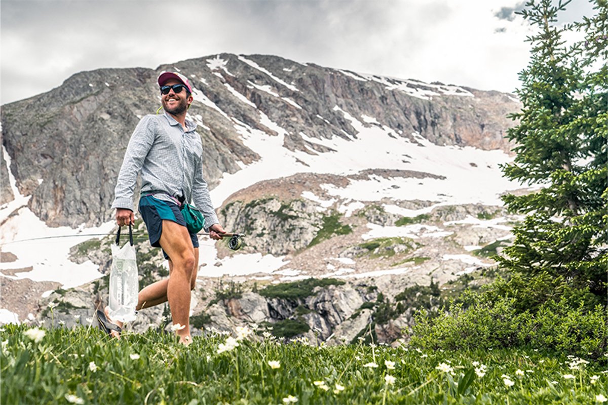 Hiker walks through an alpine meadow carrying a filled water resevoir, with rocky, snow-dusted mountains in the background.