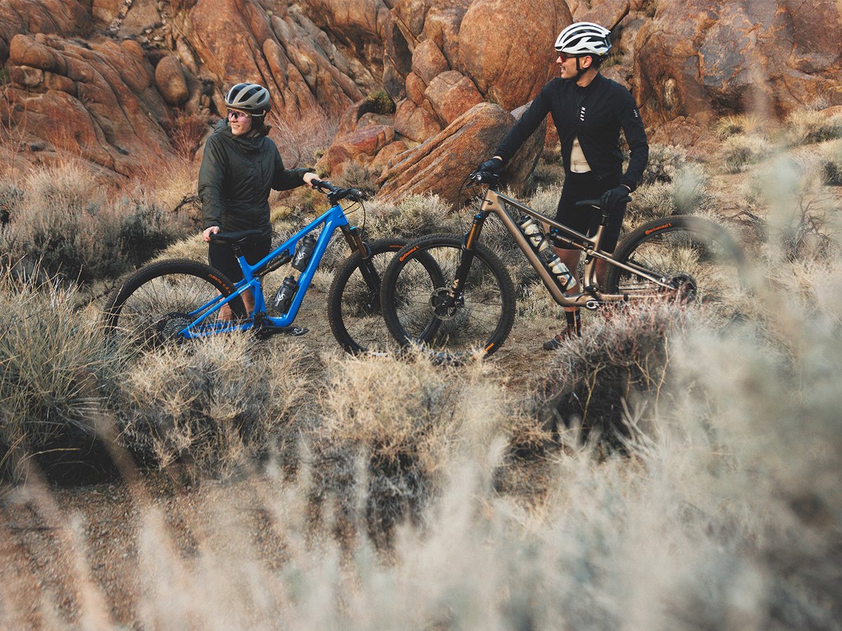 Two different height riders standing next to eachother holding their bikes with desert brush all around.