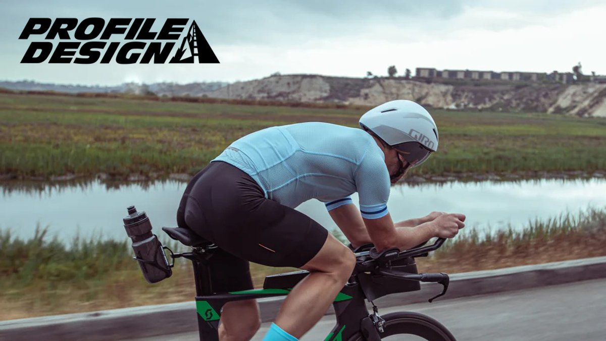 Male cyclist in aerodynamic position wearing light blue cycling jersey and white helmet on scenic rural road with wetlands landscape background.