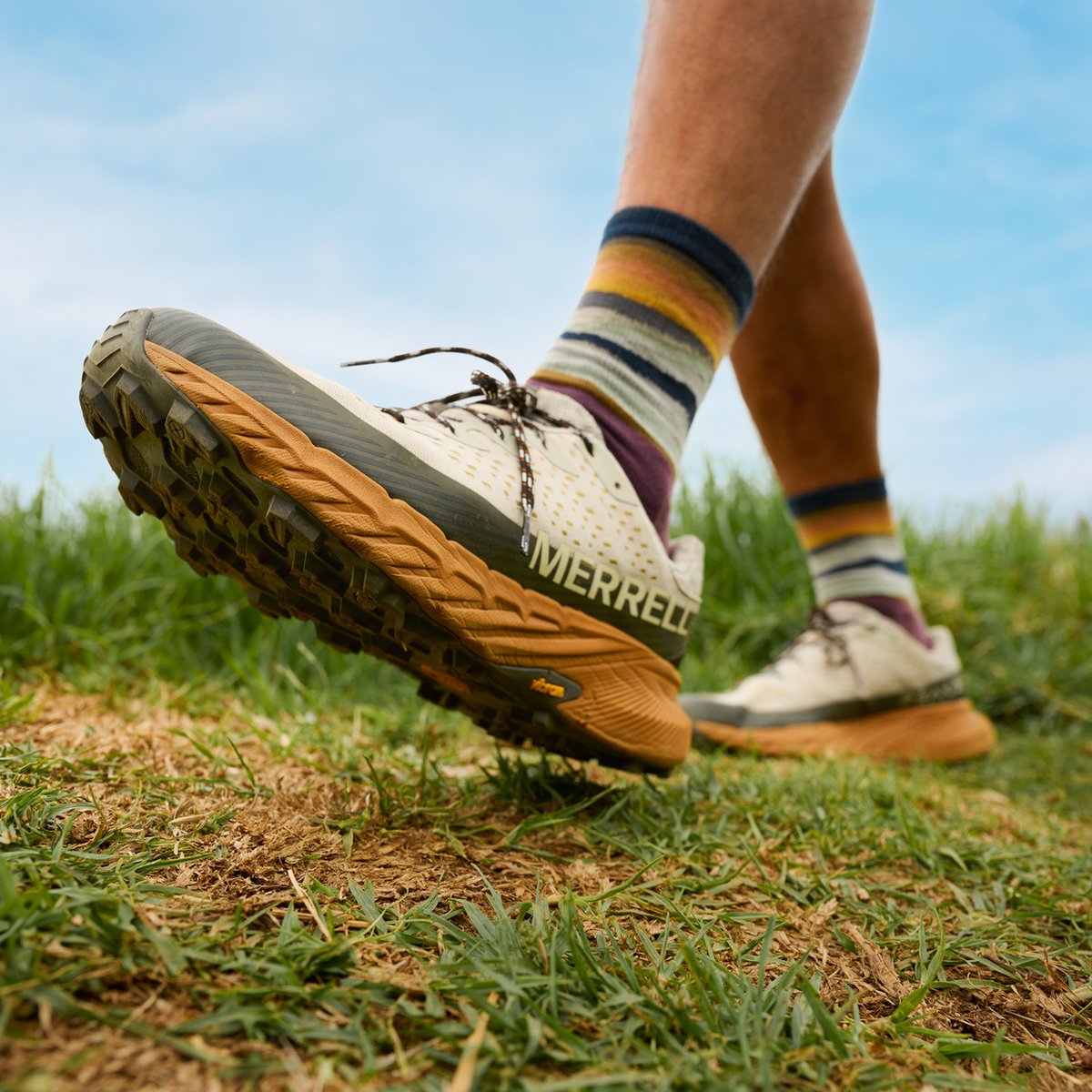 A person walks on a grassy trail wearing Merrell Agility Peak 5 trail running shoes with thick Vibram soles and striped multicolored socks, under a bright blue sky.