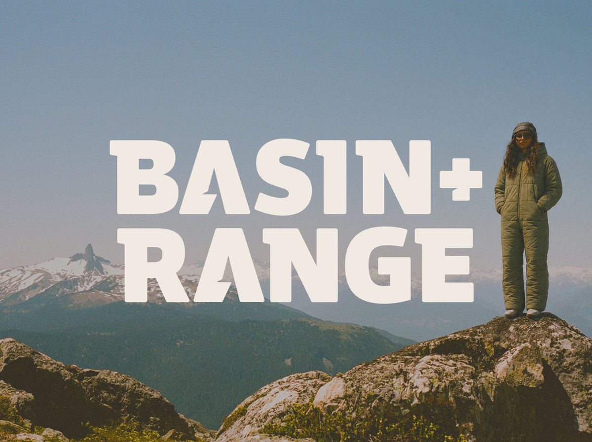 Basin+ Range logo overlaid on a person in an olive insulated one-piece suit standing on a granite summit with snow-capped peaks behind them.