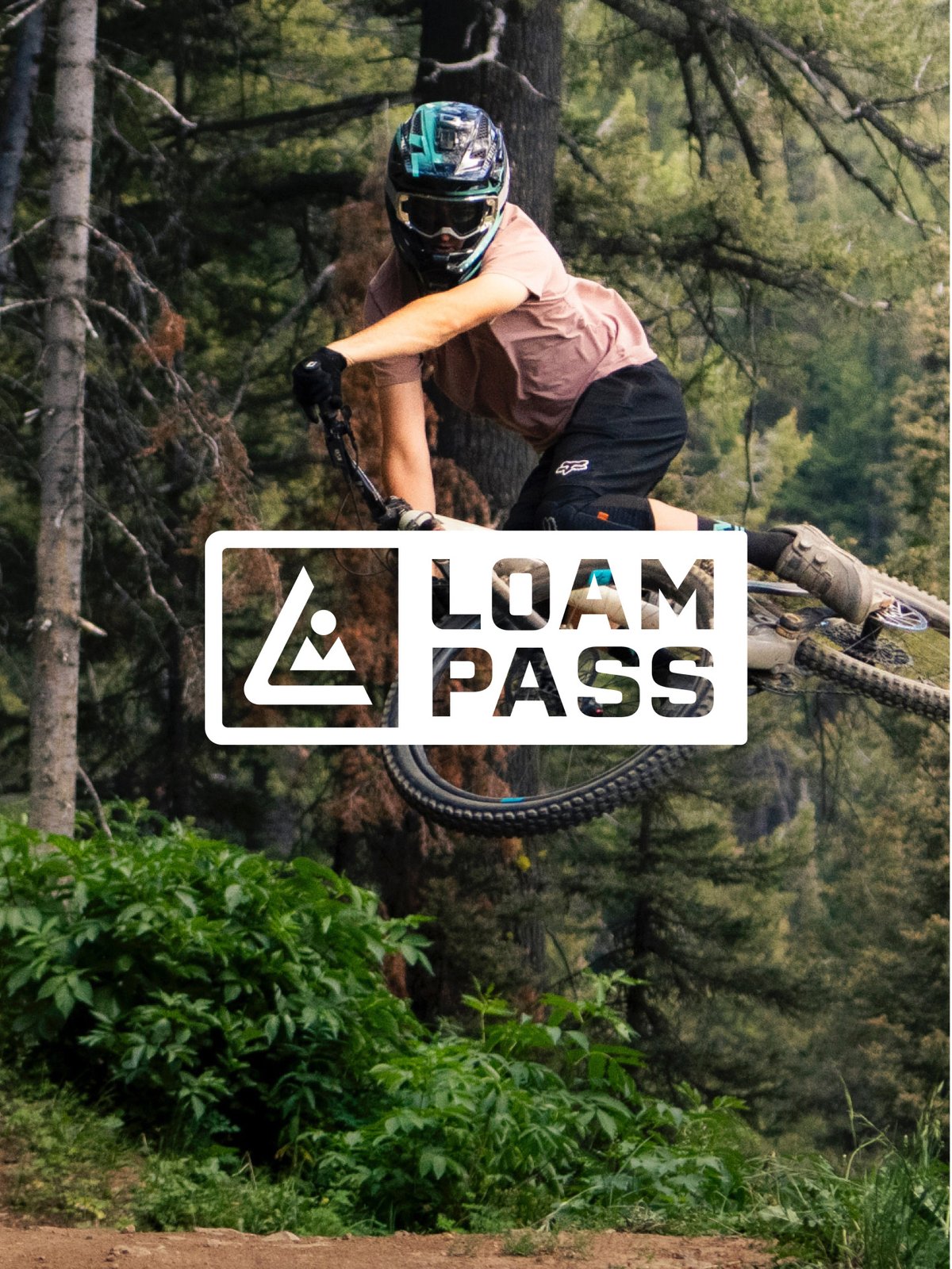 Mountain biker launching off a jump in a forested trail park, featuring the Loam Pass logo.